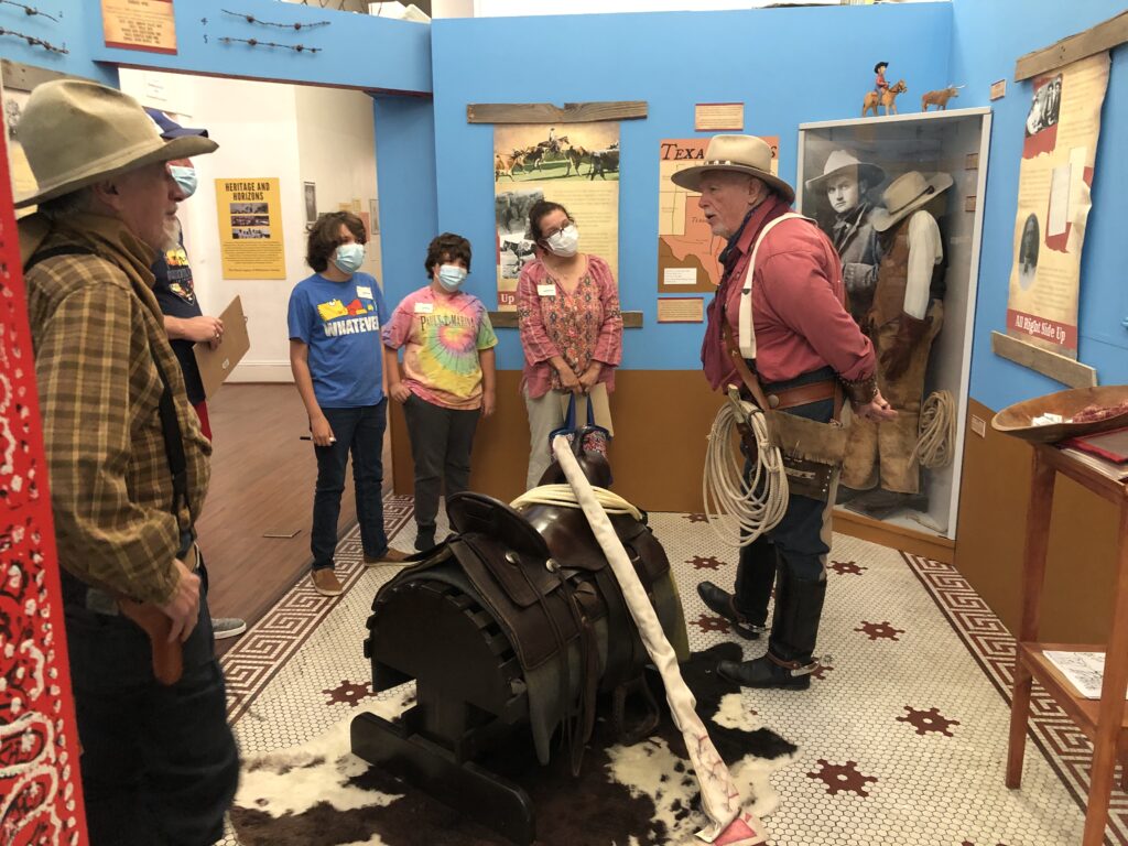 A gallery room with blue and brown walls, a fake cowskin rug over tile flooring and a saddle on a wooden mount in the middle of the exhibit. A docent dressed as a cowboy leans against the wall on the far left as another docent, also dressed as a cowboy, gives a tour to four visitors. a display case with cowboy clothing is in the far right corner, and information panels, a map, and barbed wire hang on the walls.